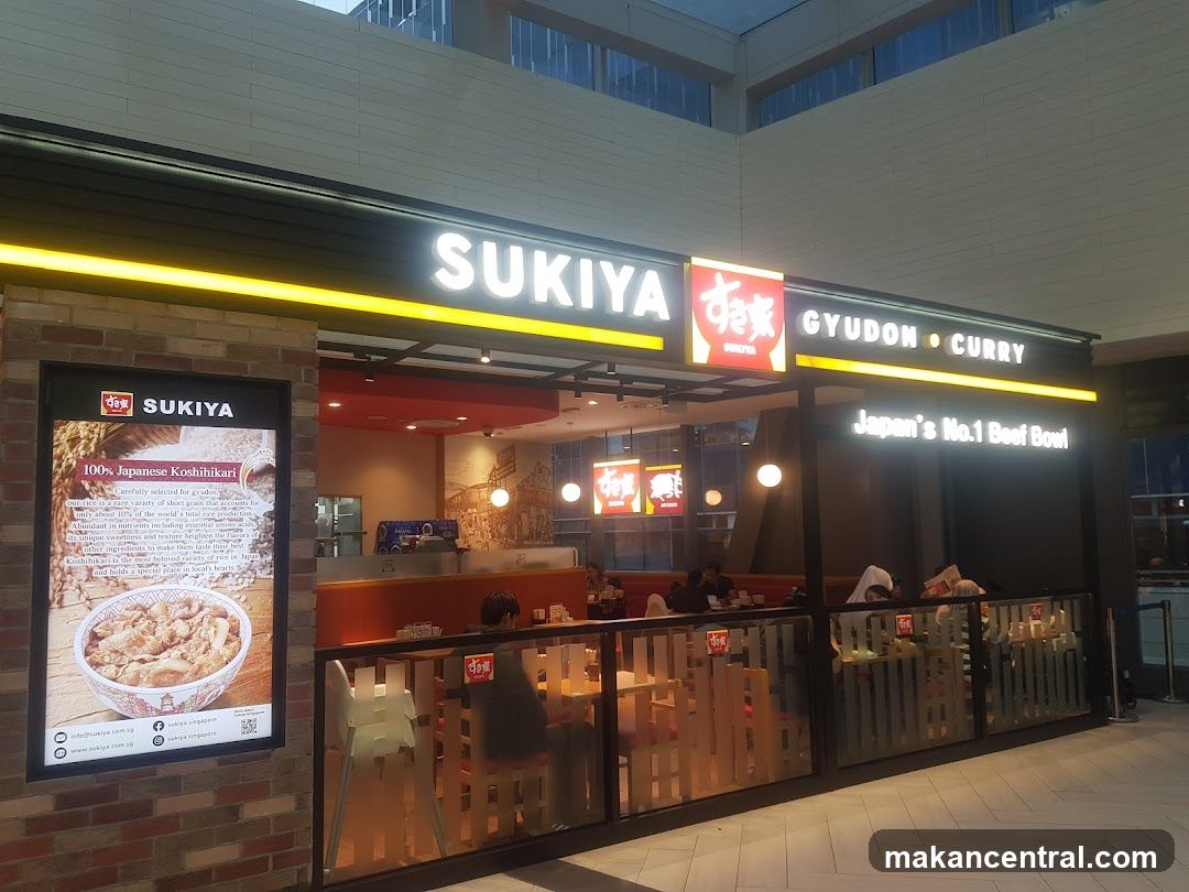 The exterior of a Sukiya restaurant, a Japanese fast-food chain. The storefront features a prominent sign with the restaurant's name, "SUKIYA," along with "GYUDON • CURRY." A large illuminated display on the left showcases "100% Japanese Koshihikari" rice and a bowl of gyudon. The restaurant interior is visible, with tables and chairs set up for dining.