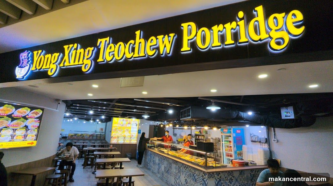 A brightly lit storefront for Yong Xing Teochew Porridge. The shop's name is displayed prominently in large yellow lettering on a black background above the entrance. Inside, tables and chairs are set up for dining, and a counter displays various food items. Staff members are visible behind the counter, serving customers. A menu board with pictures of dishes is displayed on the left side of the entrance.