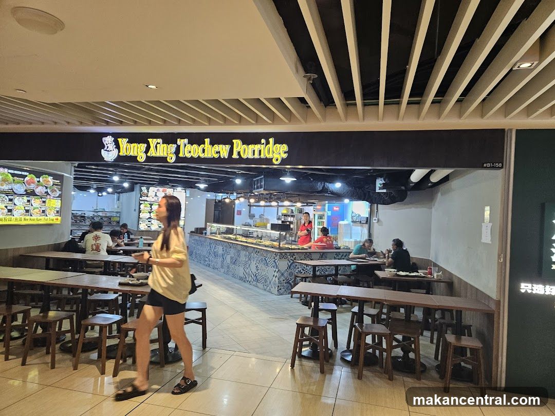 The exterior of a food stall with a prominent sign that reads "Yong Xing Teochew Porridge." Inside, customers are seated at tables, and staff members are visible behind a counter. There are also menu boards displayed with food items.
