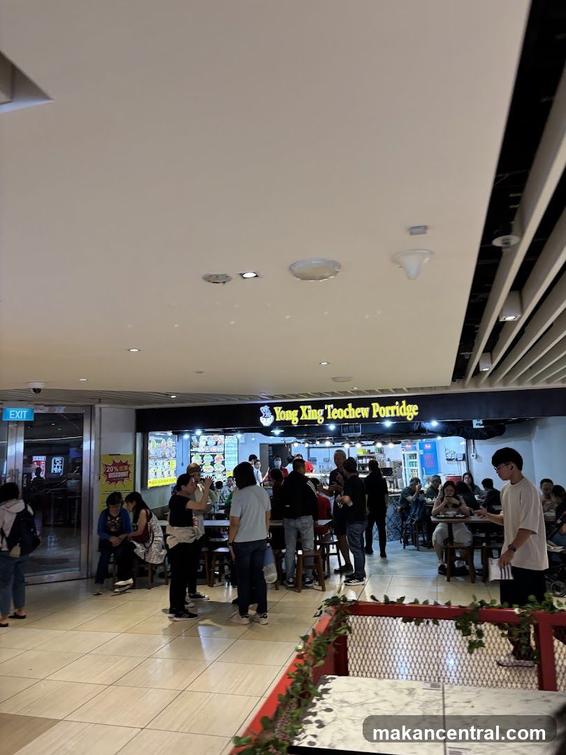 A busy food stall with customers seated at tables and standing in line. The stall has a prominent sign that reads "Yong Xing Teochew Porridge." There are menu boards displayed behind the counter, showing various food items. A sign on the left indicates a "20% discount."