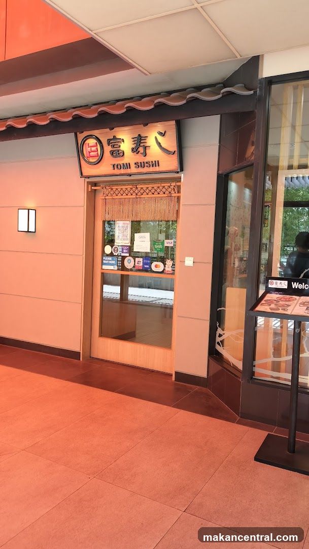 The exterior of a sushi restaurant with a wooden door and glass windows. A sign above the door reads "Tomi Sushi" in English and Japanese characters. Various payment method stickers and a "Please Wait Here" sign are visible on the door. A menu stand is placed to the right of the entrance.