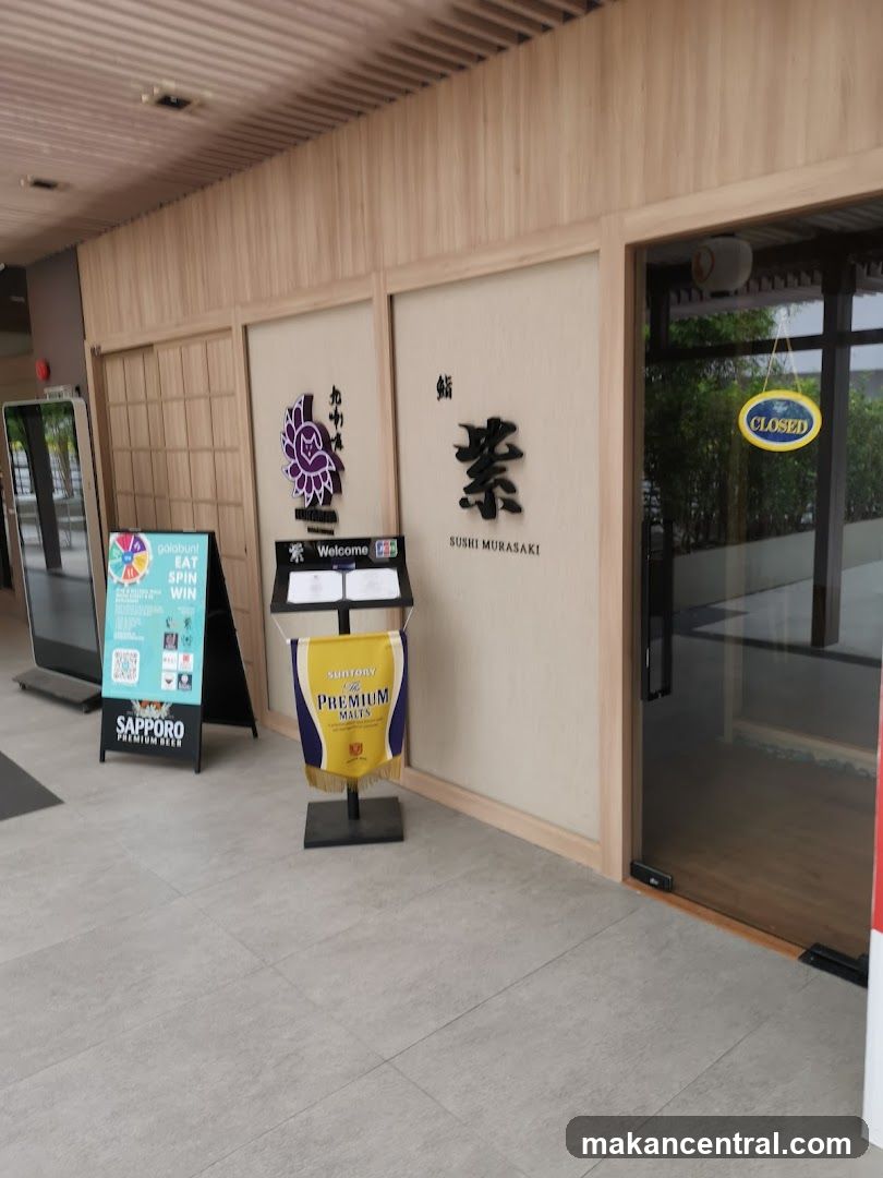 The exterior of a sushi restaurant with Japanese-style wooden paneling. A large sign with Japanese characters and "SUSHI MURASAKI" is visible on the wall. A "CLOSED" sign hangs on the glass door. Promotional signs for "galabunt" and "Sapporo Premium Beer" are placed outside. A banner for "Suntory Premium Malts" is also displayed.