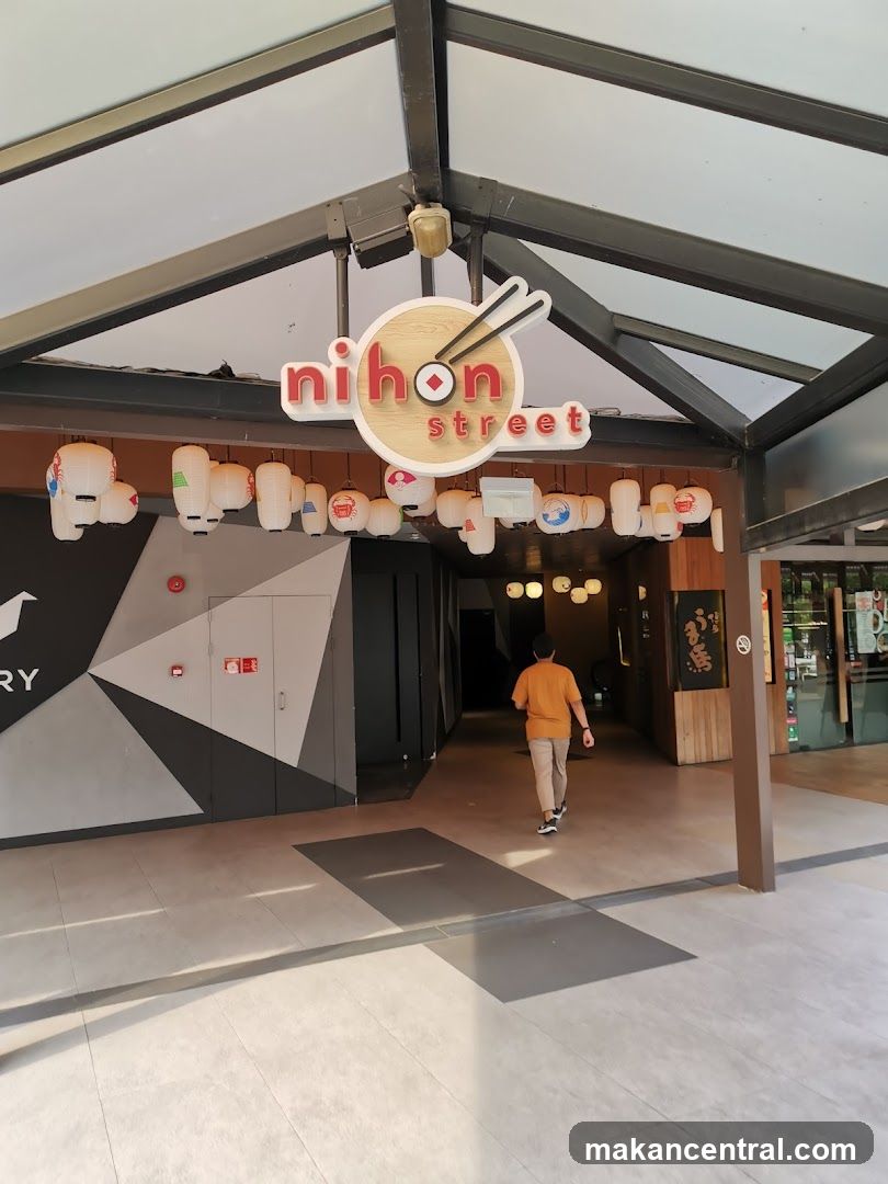 The exterior entrance of a Japanese restaurant called Nihon Street. A sign with the restaurant's name is visible above the entrance, featuring chopsticks and a red circle. Traditional Japanese lanterns hang in front of the entrance. A person is walking into the establishment.