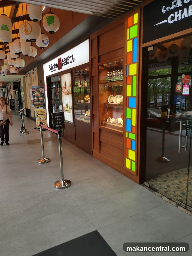 A row of Japanese restaurants with visible signage. The leftmost shop has a sign that reads 'Saboten' with Japanese characters. It features a display window showcasing food items and products. Next to it is another establishment with a wooden facade and a colorful stained-glass-like panel. To the right, a glass door leads into a restaurant with a black sign above it. A woman walks past the storefronts.