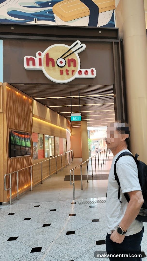 The exterior of a Japanese restaurant called Nihon Street. The restaurant has a modern design with wooden paneling and a prominent sign above the entrance. An exit sign is visible hanging from the ceiling. A person is standing in the foreground, partially obscuring the view of the entrance.