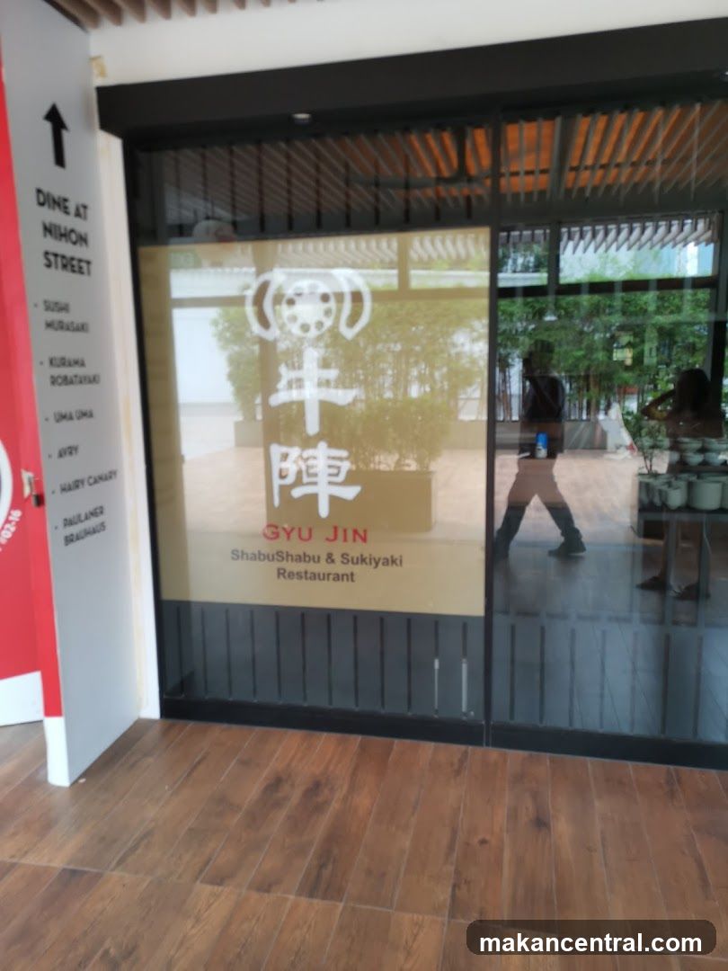 The storefront of Gyu Jin, a ShabuShabu & Sukiyaki Restaurant. The restaurant's name and logo are visible on the glass facade. To the left, a red and white sign lists various dining options, including "DINE AT NIHON STREET" and specific restaurant names.