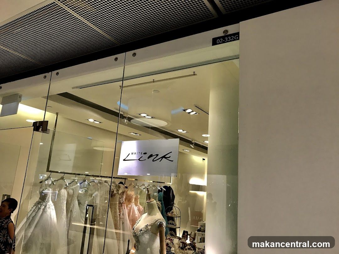 A bridal shop with a glass storefront displaying several wedding dresses on hangers and mannequins. The shop's name, "WHITE Link", is visible on a sign inside the window. A woman is partially visible on the left side of the image, looking into the shop.