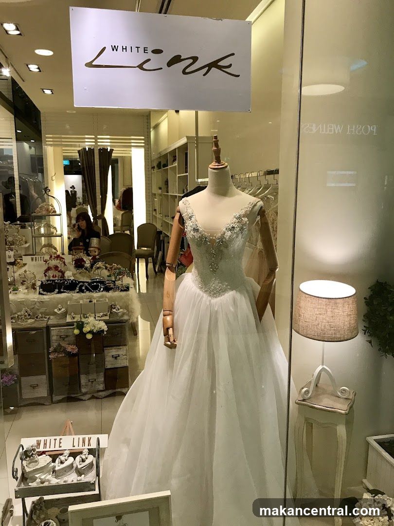 A bridal shop window display featuring a white wedding dress on a mannequin. The shop's name, "WHITE LINK," is visible on a sign above the display. The window also shows accessories and other dresses inside the shop.