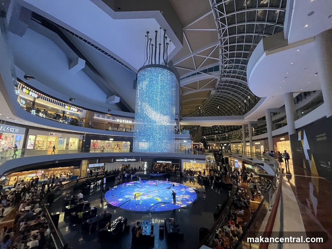 Interior of a large, modern shopping mall with a central water feature and light display.