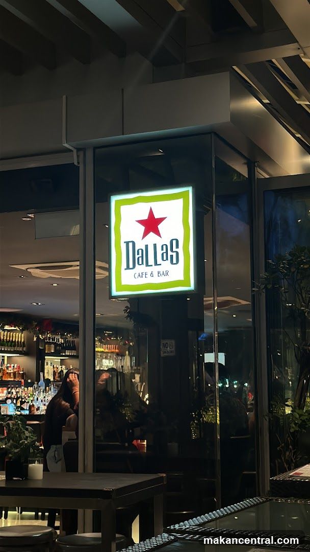 A lit sign for Dallas Cafe & Bar is displayed on the exterior of the building. The sign features a red star above the word "Dallas" in green lettering, with "CAFE & BAR" below it, all set against a white background with a green border. Through the glass windows, a bar area with shelves of bottles and a person can be seen.