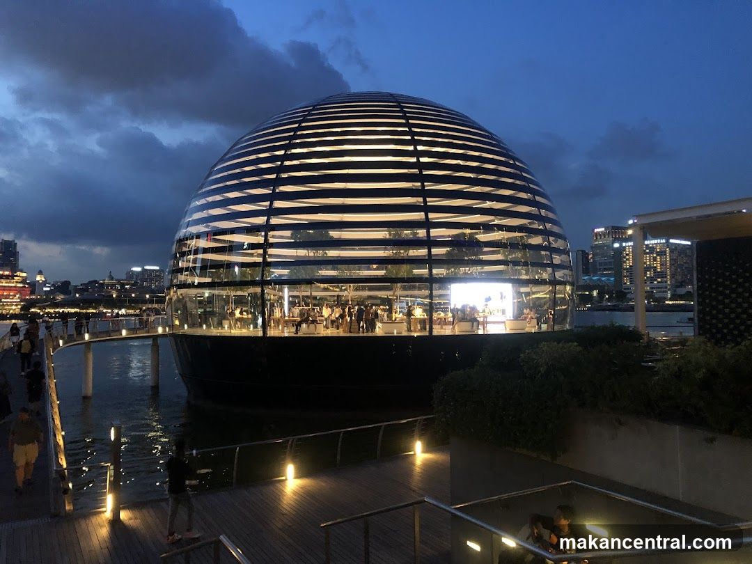 A unique spherical Apple Store with a glass facade, illuminated from within, sits on the water at dusk. A walkway leads to the entrance, with people visible inside the store. The surrounding cityscape and water are reflected in the building's exterior.