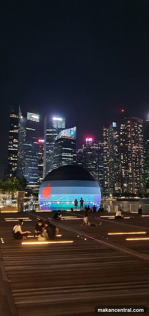 A nighttime exterior view of a spherical Apple Store with its logo illuminated on the facade. The store is situated in a waterfront area with a modern cityscape in the background. People are seen sitting and relaxing on a wooden deck in the foreground.
