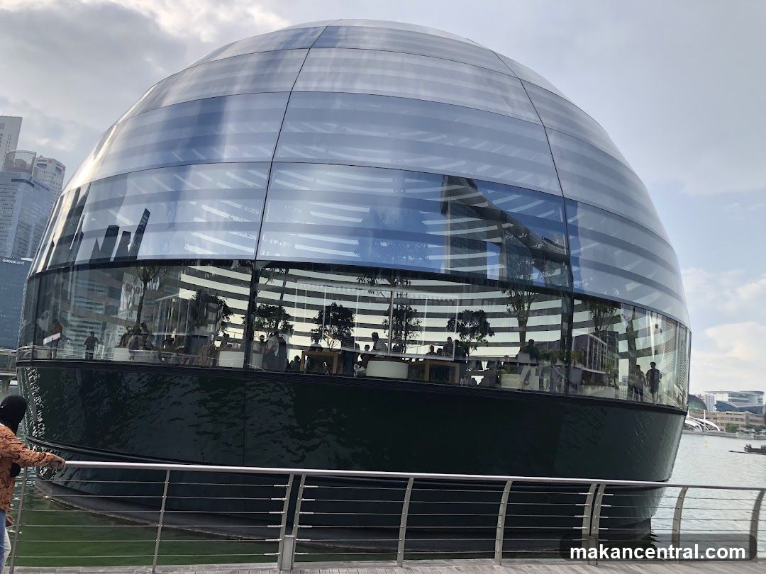 A spherical glass building with a dark base, situated on the water. The building has a modern, futuristic design. Inside, people can be seen browsing and interacting with displays. The exterior reflects the surrounding cityscape and sky.
