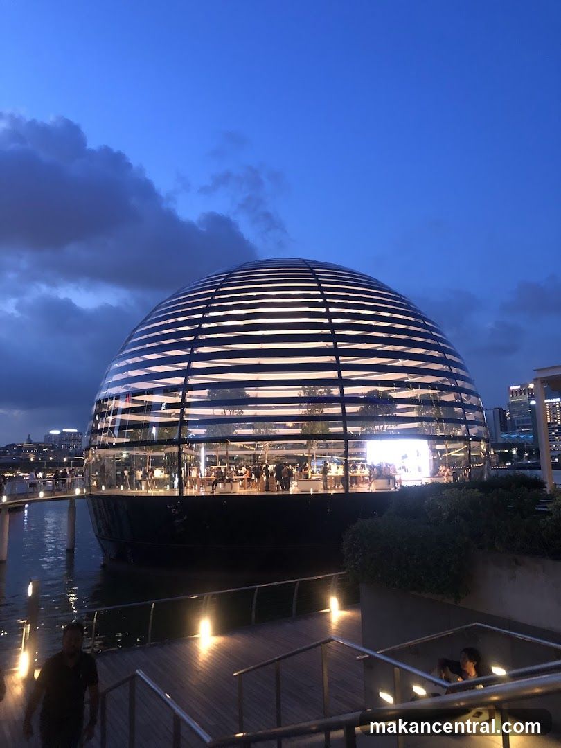 A modern, spherical glass building with horizontal white accents, situated on the water's edge. The interior is brightly lit, revealing people browsing and displays of products. The exterior is illuminated by internal lights and external pathway lights. The sky is a deep blue with some clouds.