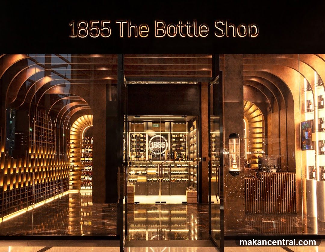 The exterior of a modern bottle shop with a neon sign that reads "1855 The Bottle Shop". The entrance features large glass doors and windows, revealing a well-lit interior with extensive wine displays. The shop's interior design includes arched shelving units illuminated with warm lighting, creating a luxurious ambiance.