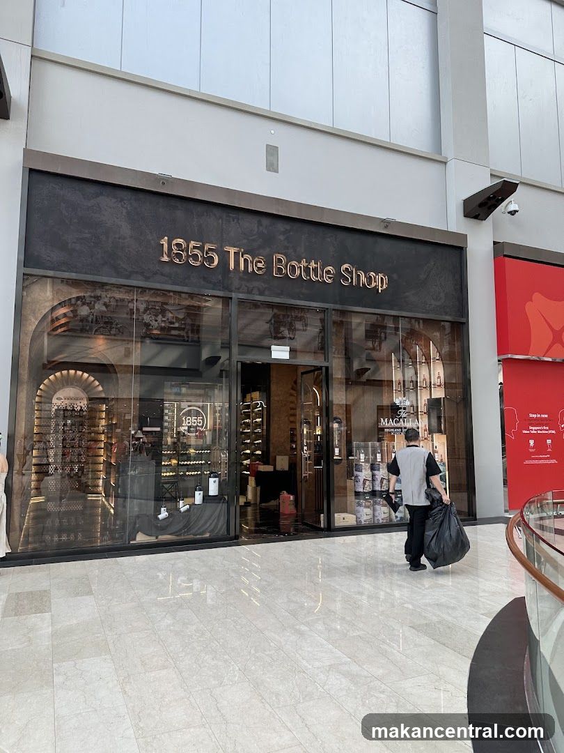 The exterior of a bottle shop with the name "1855 The Bottle Shop" displayed prominently above the entrance. The shop has large glass windows showcasing shelves stocked with bottles and displays inside. A person is seen walking away from the shop carrying a large black trash bag.